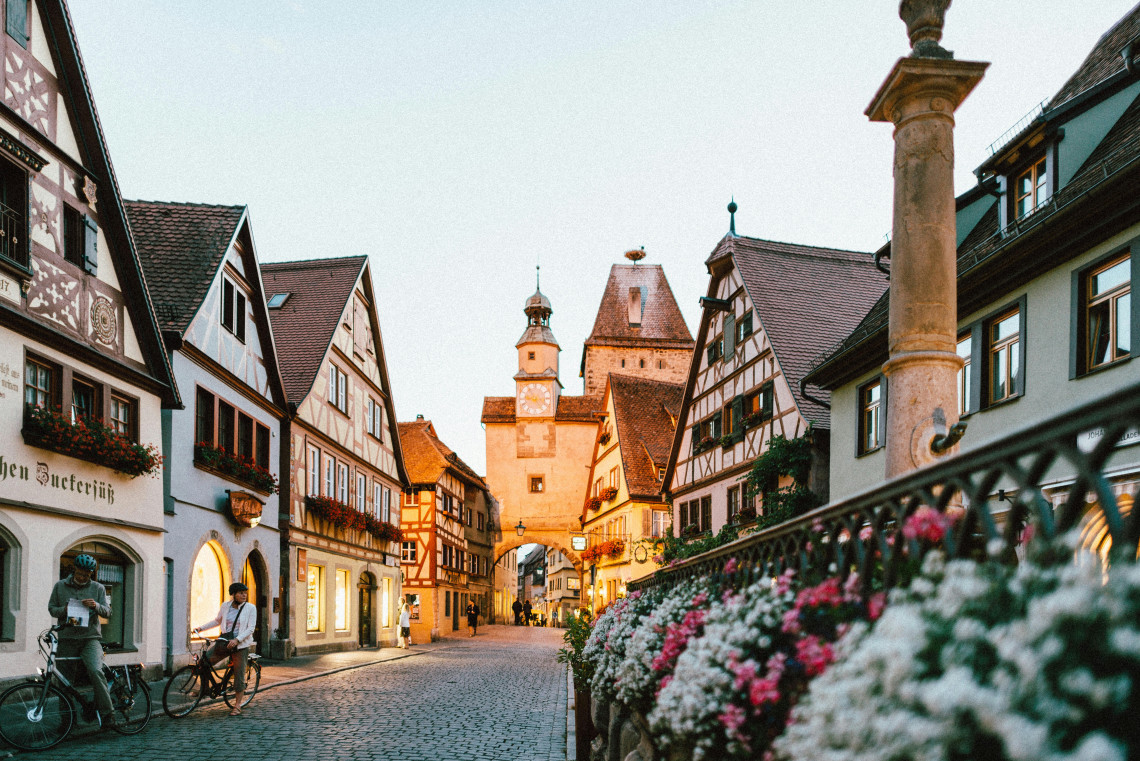 A cobblestone street in the historic centre of Rothenburg ob der Tauber, Germany, lined with colourful half-timbered houses and traditional shops. A medieval tower rises at the end of the street, while flower-filled railings frame the foreground.