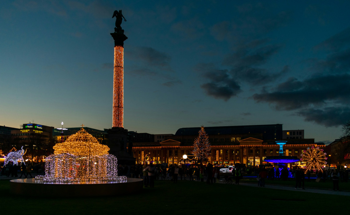Evening view of the Stuttgart Christmas Market at Schlossplatz in Stuttgart, Germany, decorated with illuminated festive displays and Christmas trees. The Jubilee Column rises above the square against the dark winter sky.