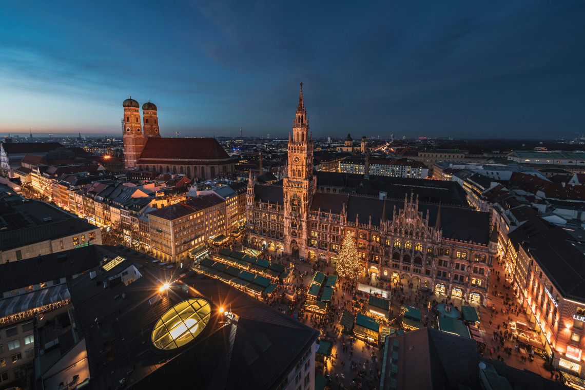 Aerial evening view of the Christmas market at Marienplatz in Munich, Germany, surrounded by illuminated historic buildings. The New Town Hall stands beside festive market stalls, with the twin towers of the Frauenkirche visible in the background.