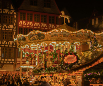 Close-up of an ornate illuminated fairground carousel at a Christmas market at night, with a golden horse on top, warm glowing lights, decorated market stalls below, and half-timbered buildings in the background