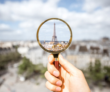 Hand holding a magnifying glass with the Eiffel Tower reflected inside against the Paris skyline, France travel decision concept