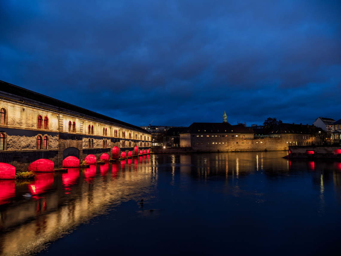 Night view of Barrage Vauban in Strasbourg, France, illuminated beside the River Ill. The historic 17th-century defensive bridge reflects on the calm water near the Ponts Couverts in the city's UNESCO-listed historic centre.
