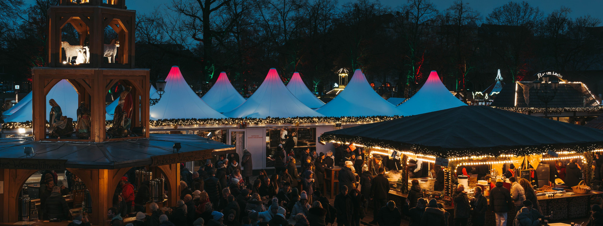 Evening crowds gather between illuminated stalls at a Christmas market in Budapest, Hungary. Strings of warm lights, decorated wooden structures and white peaked tents fill the seasonal market.