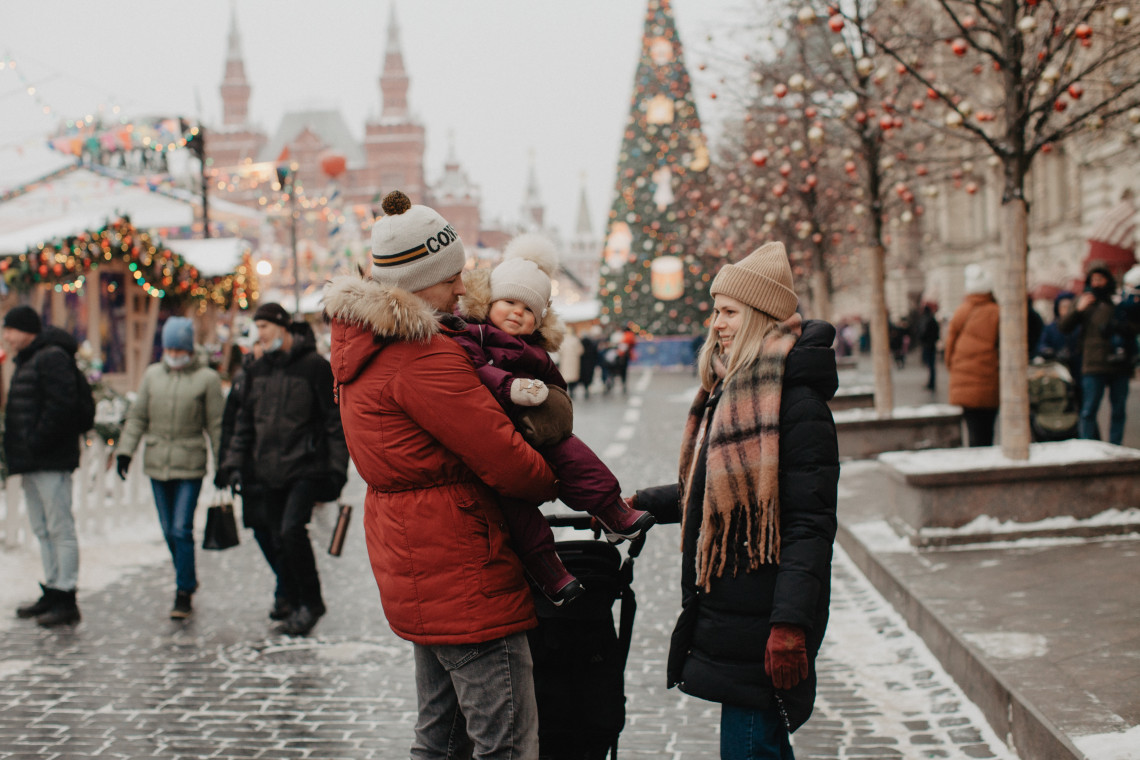 A family walks through the snow-covered Christmas market in Red Square, Moscow, Russia. Decorated stalls, a large Christmas tree and the Kremlin towers appear in the background.
