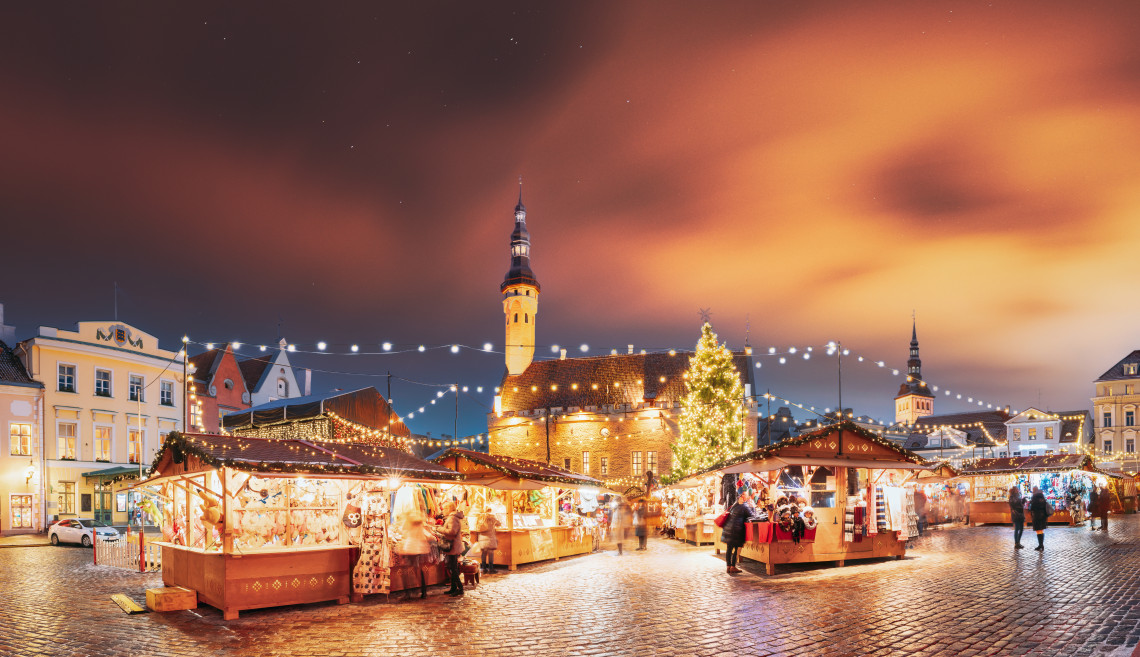Wooden stalls and a decorated Christmas tree illuminate Tallinn Christmas Market in Tallinn, Estonia. The medieval Town Hall overlooks the cobbled square beneath an orange evening sky.