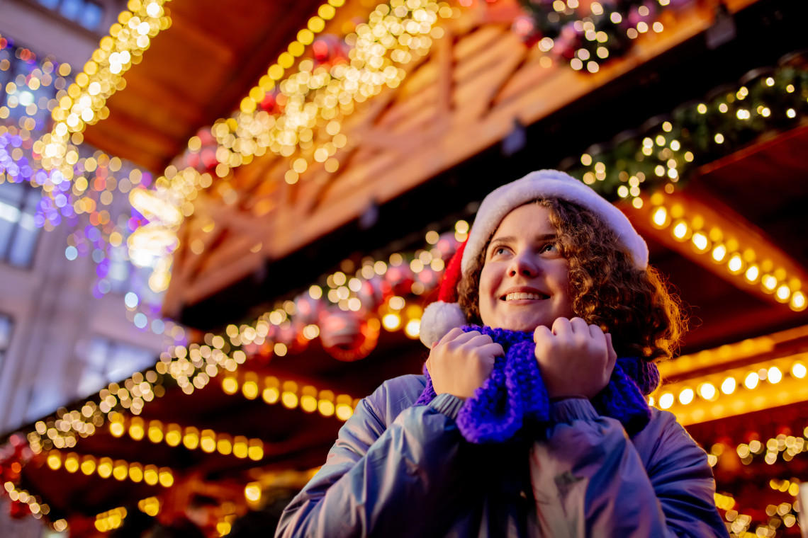 A visitor wearing winter clothing stands beneath warm seasonal lights at Tallinn Christmas Market in Tallinn, Estonia. Decorated wooden stalls and hanging ornaments create a softly blurred background.