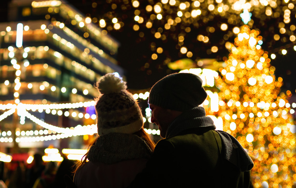 Two visitors view the seasonal lights and illuminated Christmas tree at a market in Tallinn, Estonia. Strings of warm lights and decorated stalls fill the softly focused evening scene.