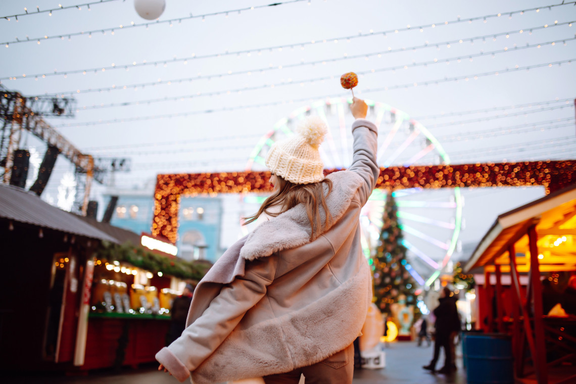 A visitor holds a colourful lollipop above the wooden stalls of a Christmas market in Budapest, Hungary. An illuminated Ferris wheel, seasonal lights and a decorated tree rise in the background.