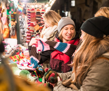 Visitors browse knitted scarves, hats and handcrafted gifts at a Christmas market stall in Budapest, Hungary. Three women examine a colourful winter scarf surrounded by seasonal merchandise.