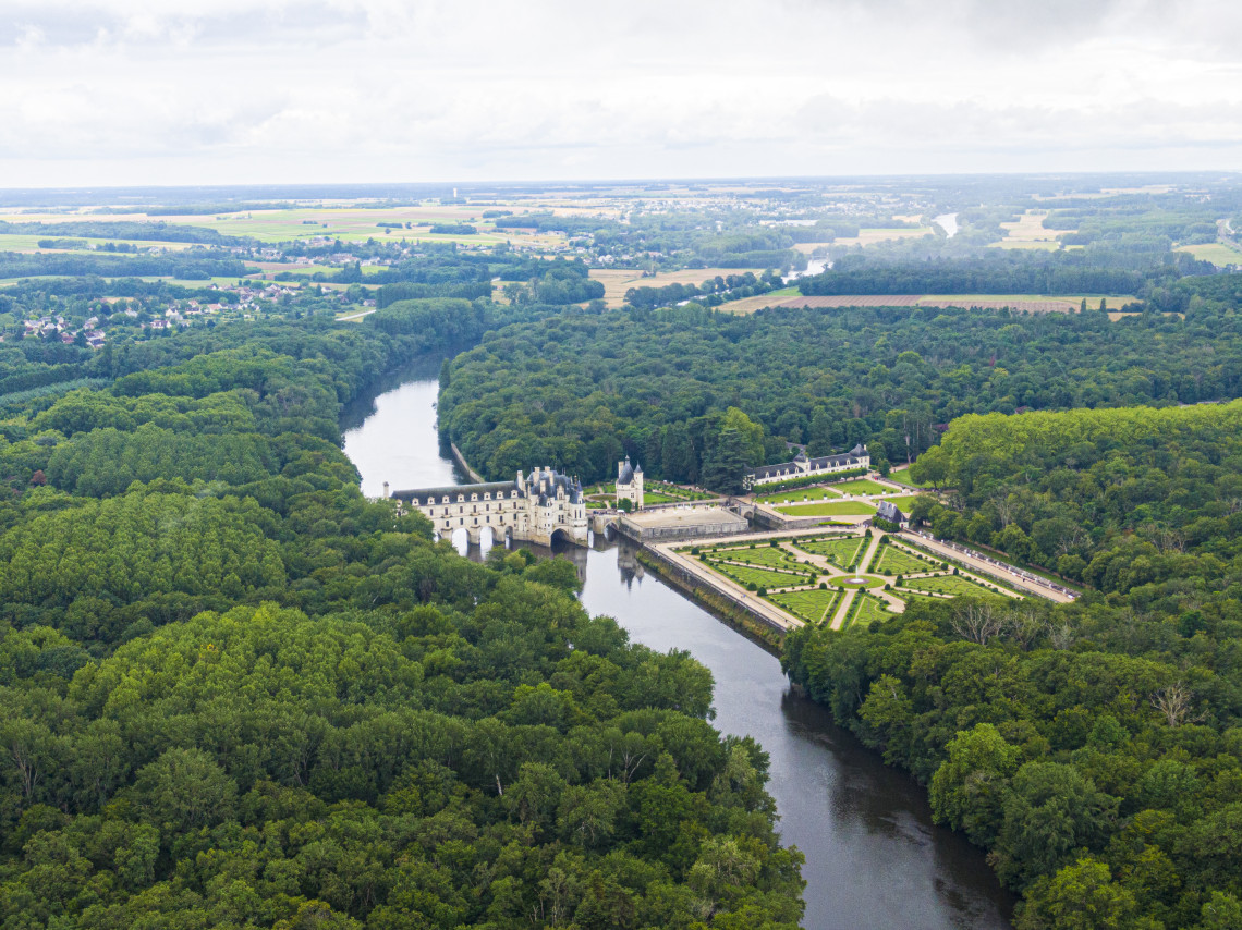 Aerial view of Chateau de Chenonceau spanning the Cher River surrounded by dense forest and formal gardens, Loire Valley, France