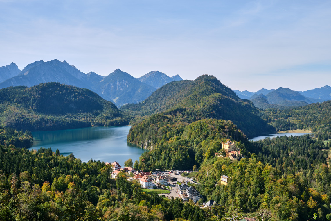 Panoramic Alpine landscape around Hohenschwangau, with Hohenschwangau Castle on a wooded hill between the Alpsee and surrounding mountains.