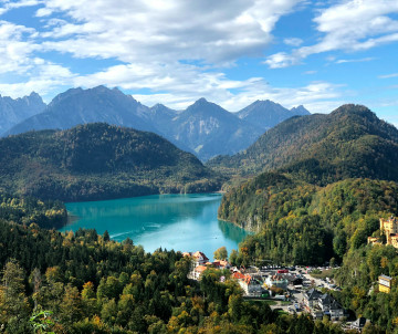 Panoramic view over Alpsee and Hohenschwangau in the Bavarian Alps, with forested hills, mountain peaks and Hohenschwangau Castle overlooking the landscape.