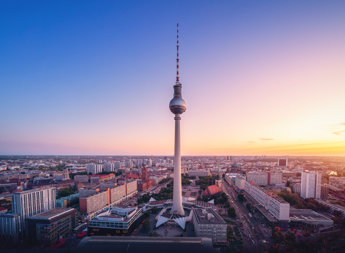 Aerial view of the Berlin TV Tower rising above the city skyline during sunset, with streets and buildings extending across Berlin.