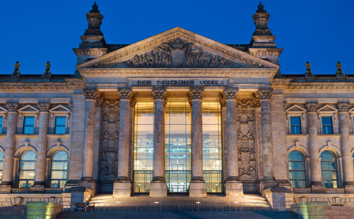 Illuminated fa&ccedil;ade of the Reichstag Building in Berlin, featuring its grand columns, stone reliefs and historic inscription.