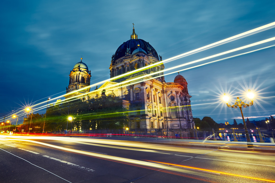Berlin Cathedral illuminated at night, with long vehicle light trails passing along the street in front of the landmark.