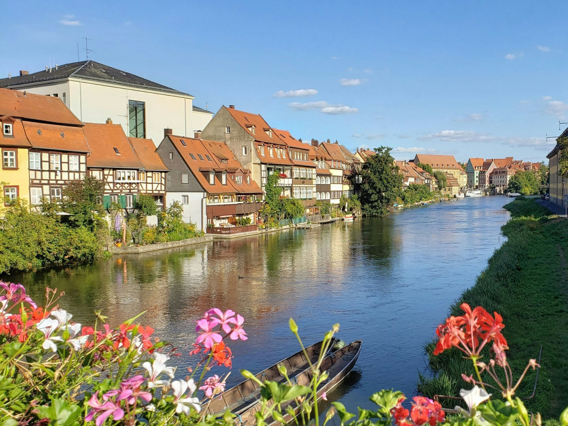 Historic half-timbered houses line the Regnitz River in Bamberg, Germany, with colourful flowers and a small wooden boat in the foreground. The waterfront district is known as Little Venice.