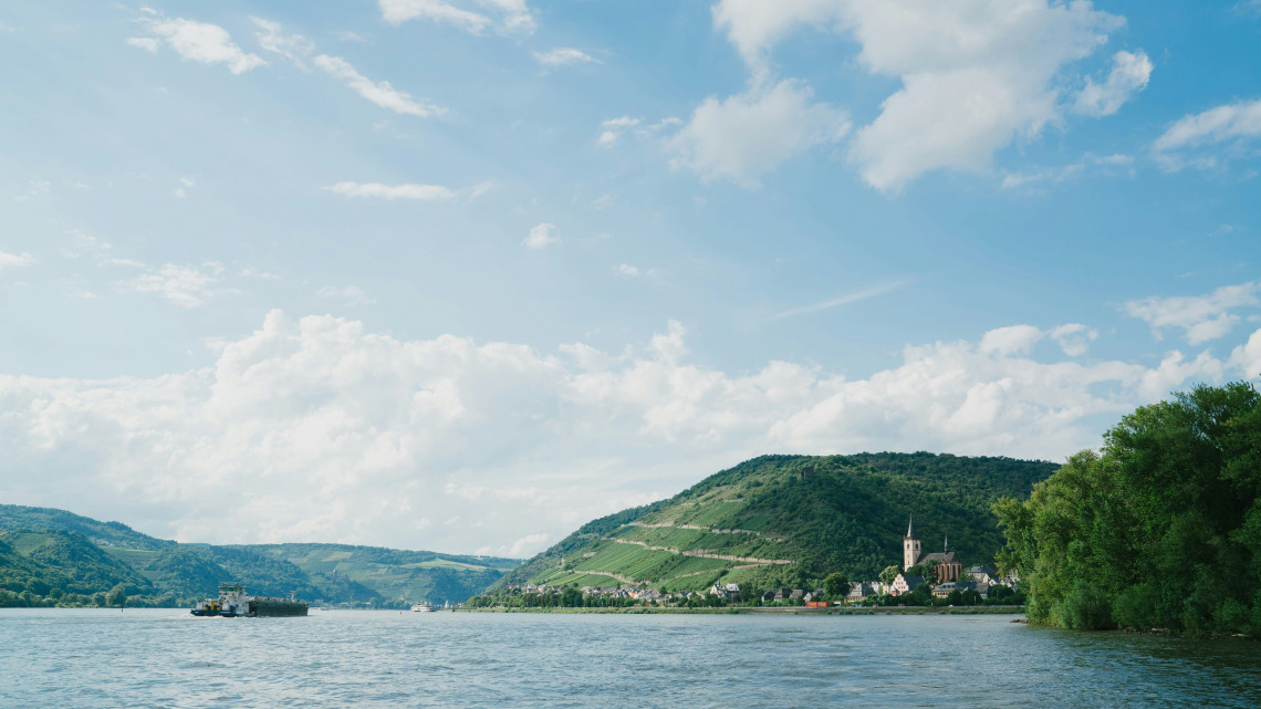 The Danube River flows through Austria&rsquo;s Wachau Valley beside a small village and vineyard-covered hills. A river vessel crosses the water beneath a bright, cloud-filled sky.