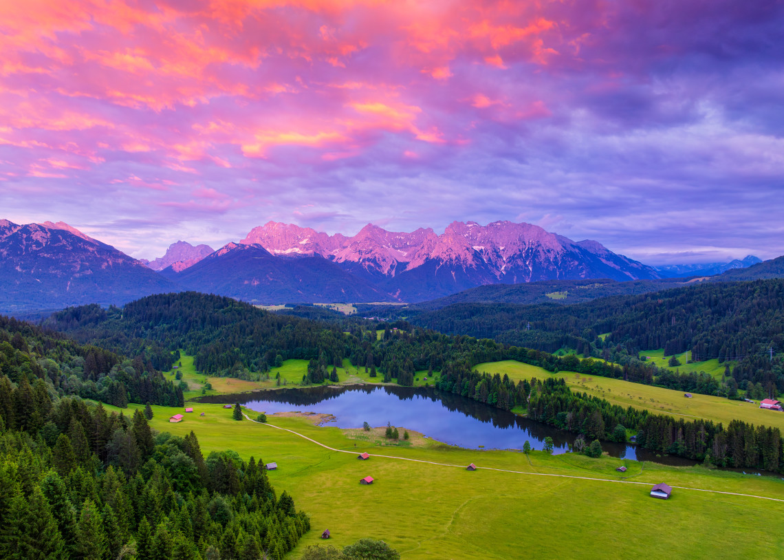 Geroldsee near Kr&uuml;n, Germany, lies among green meadows and scattered mountain huts beneath the Karwendel Alps. Pink and purple sunset clouds illuminate the Bavarian landscape.