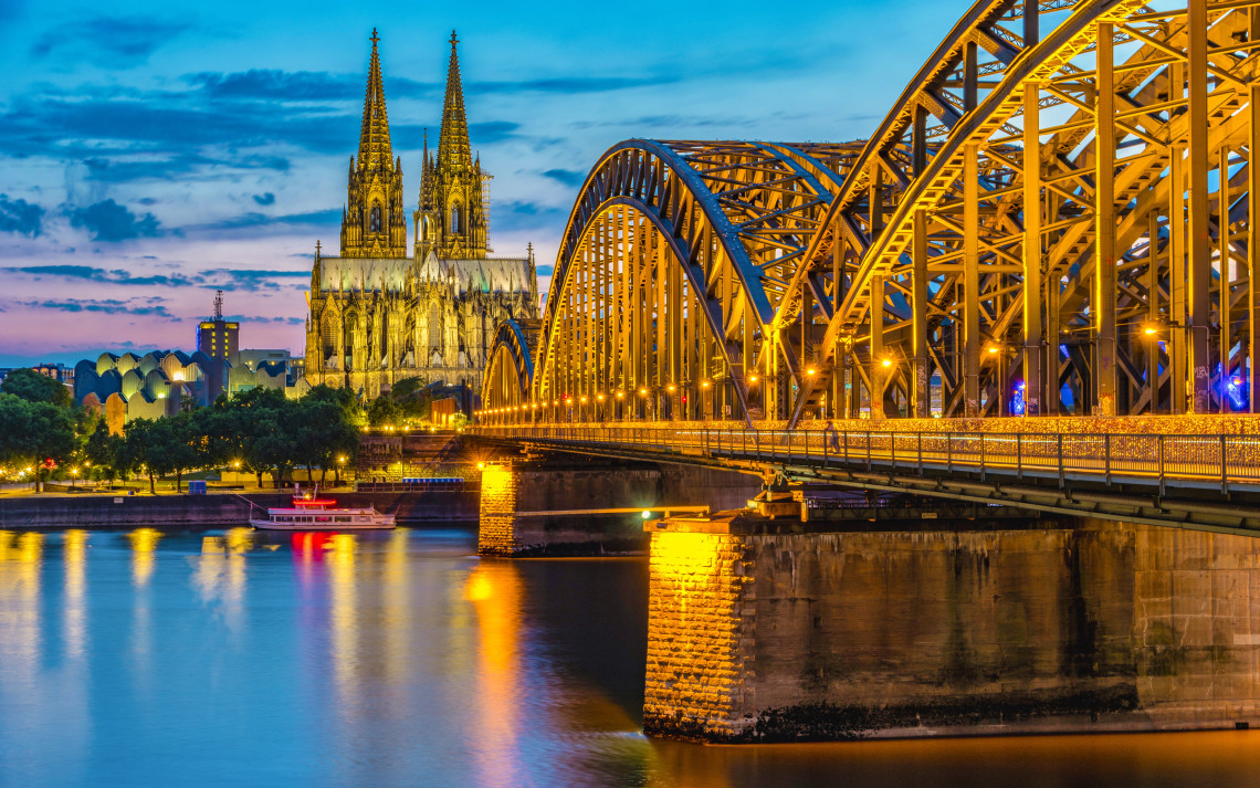 The illuminated Hohenzollern Bridge crosses the Rhine River toward Cologne Cathedral in Cologne, Germany. The Gothic cathedral towers and steel railway arches stand against the evening sky.