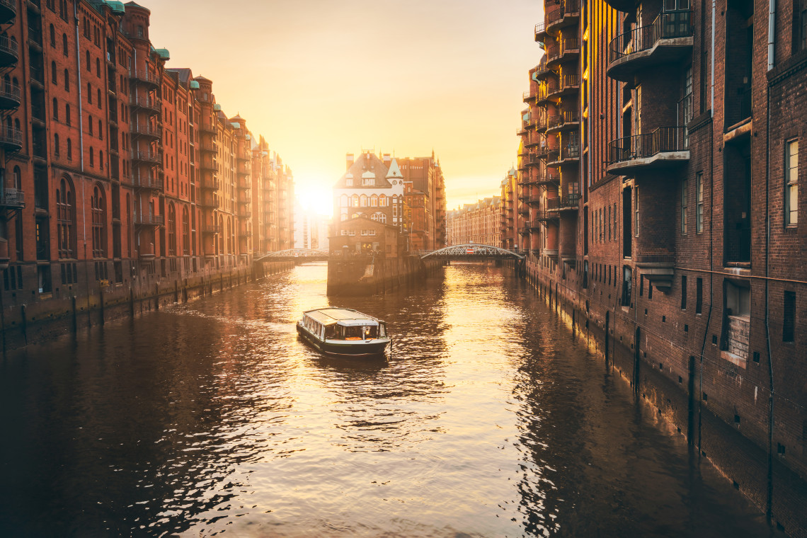 A tour boat travels through a canal in Hamburg&rsquo;s Speicherstadt district, Germany, between rows of historic red-brick warehouses. Warm sunset light reflects across the water and surrounding fa&ccedil;ades.