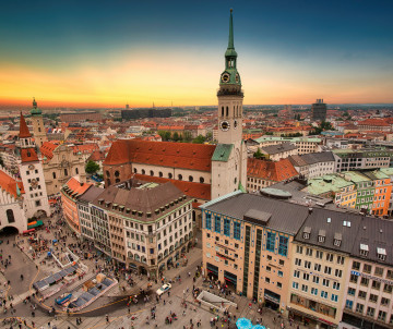 Aerial view of St Peter’s Church and Marienplatz in Munich, Germany, surrounded by red-roofed buildings, pedestrian squares and the historic city centre at sunset.