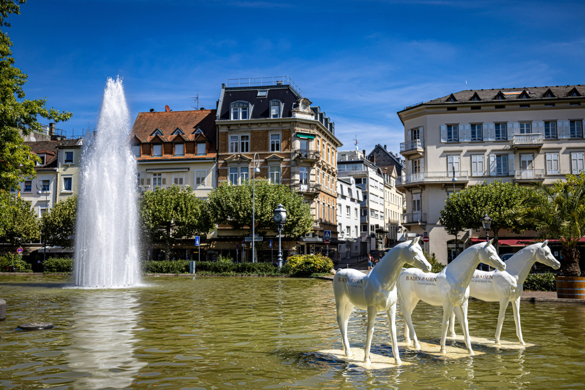 White horse sculptures stand within a large fountain in central Baden-Baden, Germany. Historic buildings, trimmed trees and a tall water jet surround the public square.