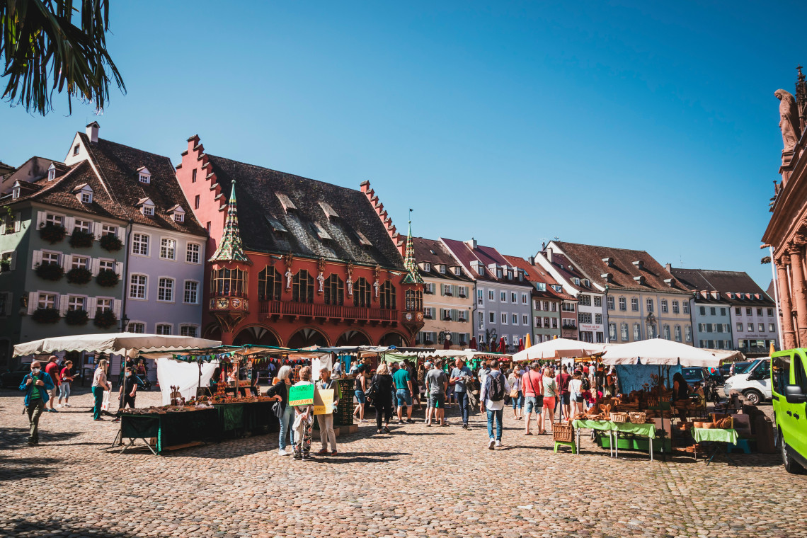 Market stalls and visitors fill M&uuml;nsterplatz in Freiburg im Breisgau, Germany. The red fa&ccedil;ade of the Historic Merchants&rsquo; Hall and surrounding Old Town buildings overlook the cobblestone square.