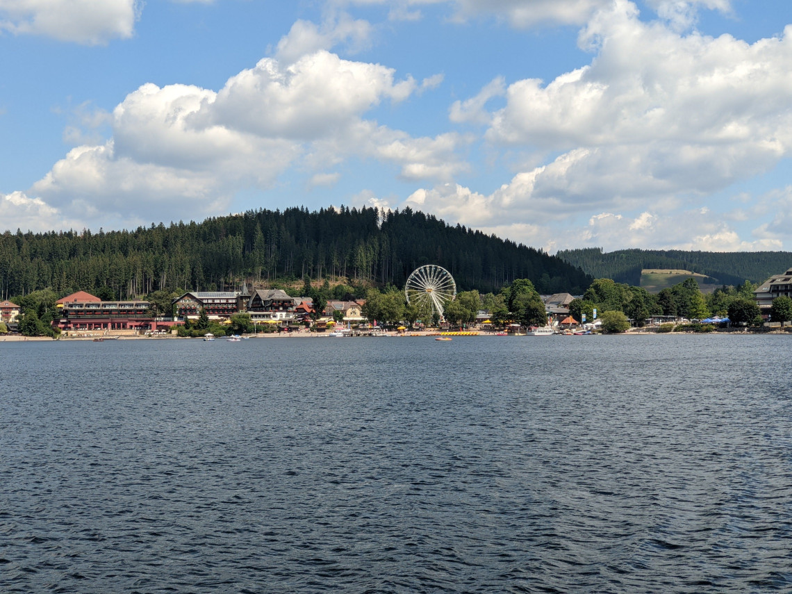 Lake Titisee stretches toward the waterfront village of Titisee-Neustadt, Germany. Hotels, trees and a Ferris wheel stand along the shore beneath forested Black Forest hills.