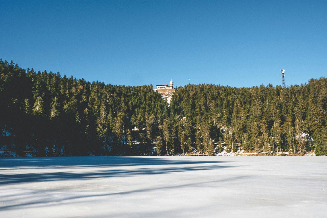 The frozen surface of Mummelsee lies beneath a densely forested hillside near Seebach, Germany. Snow covers parts of the shoreline, while buildings and a communications tower appear above the trees.