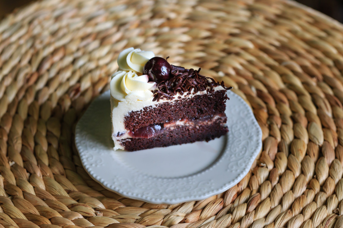 A slice of Black Forest cake is served on a white plate in Triberg im Schwarzwald, Germany. Layers of chocolate sponge, cherry filling and cream are topped with chocolate shavings and a dark cherry.