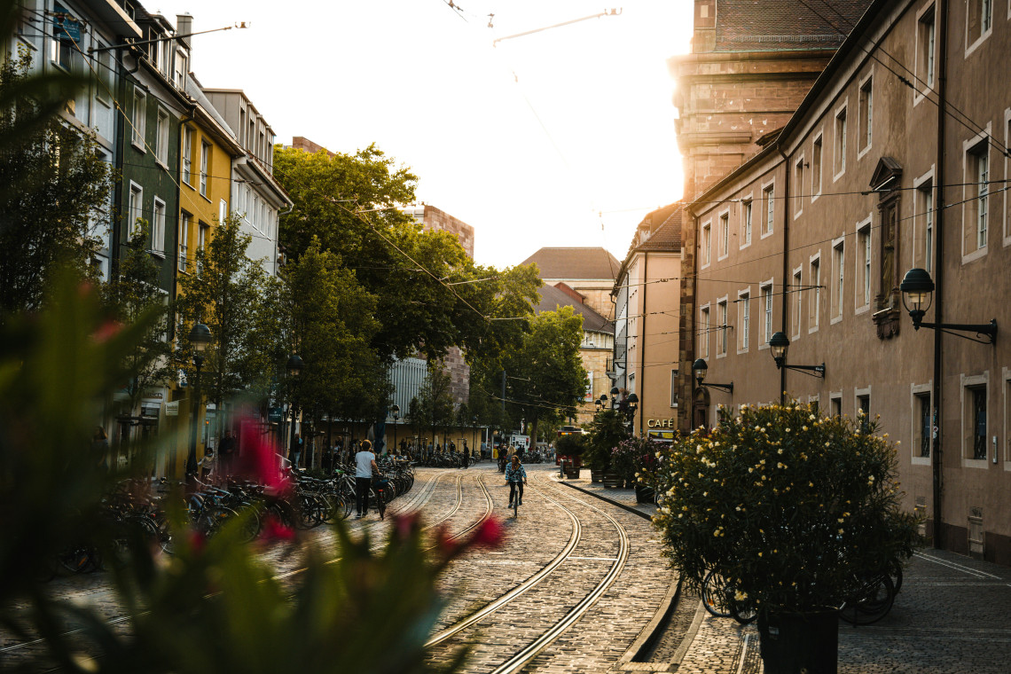 Tram tracks curve through a cobblestone street in Freiburg im Breisgau, Germany. Historic buildings, bicycles, trees and outdoor caf&eacute;s line the sunlit Old Town route.