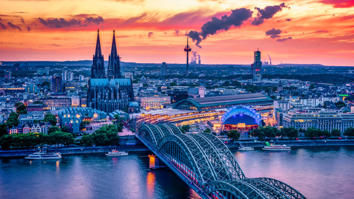 Panoramic evening view across the Rhine toward Cologne Cathedral, Cologne Central Station and the Hohenzollern Bridge beneath a colorful sunset sky.