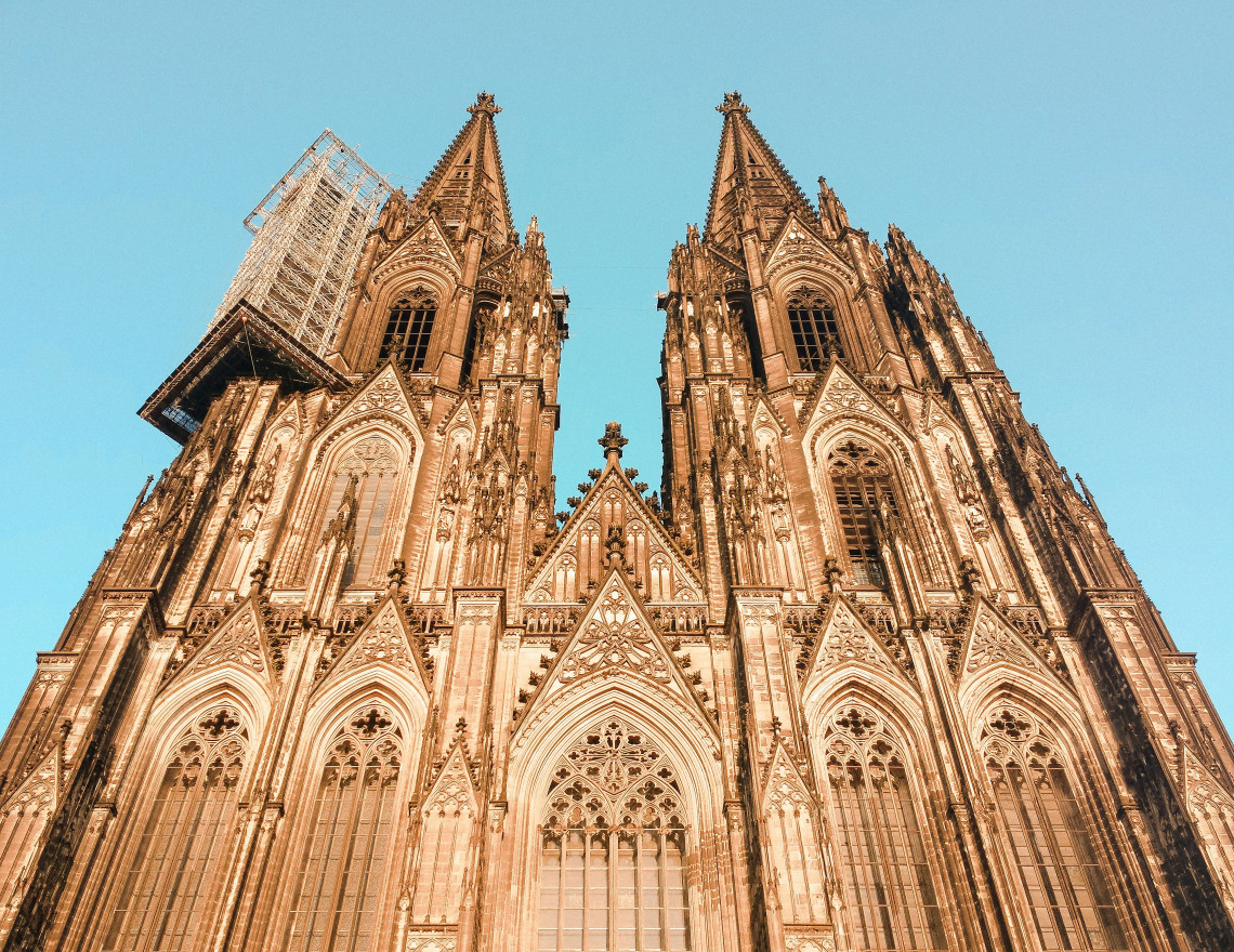 Low-angle view of the ornate Gothic fa&ccedil;ade and twin towers of Cologne Cathedral against a clear blue sky.