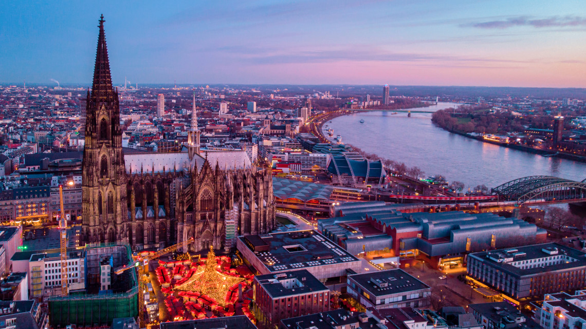 Elevated evening view of Cologne Cathedral and the surrounding city center, with an illuminated Christmas market and the Rhine River nearby.