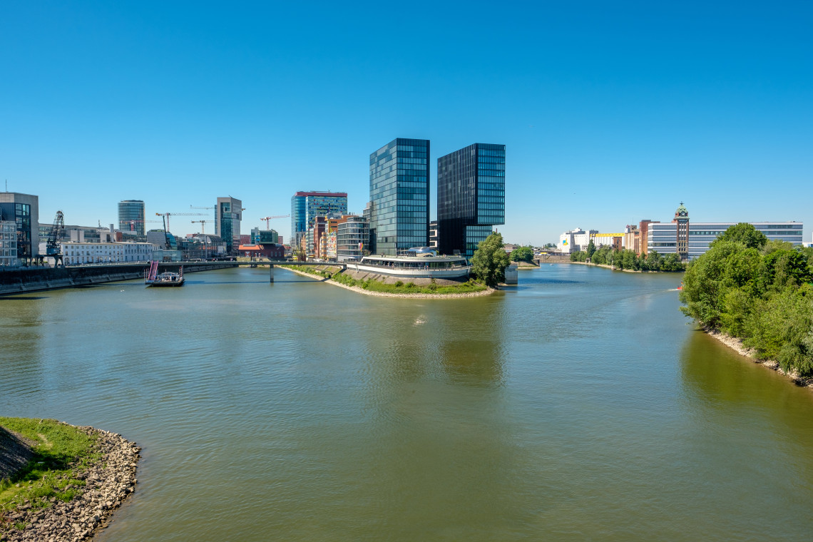 Modern office towers and waterfront architecture stand beside the Rhine at D&uuml;sseldorf&rsquo;s MedienHafen district under a clear blue sky.