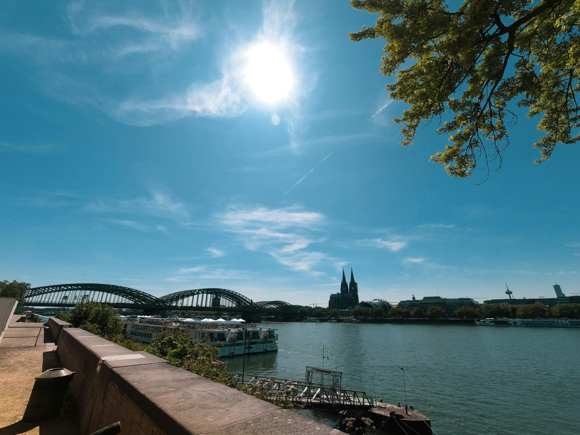Sunny view across the Rhine toward Cologne Cathedral and the Hohenzollern Bridge, with riverboats along the waterfront.