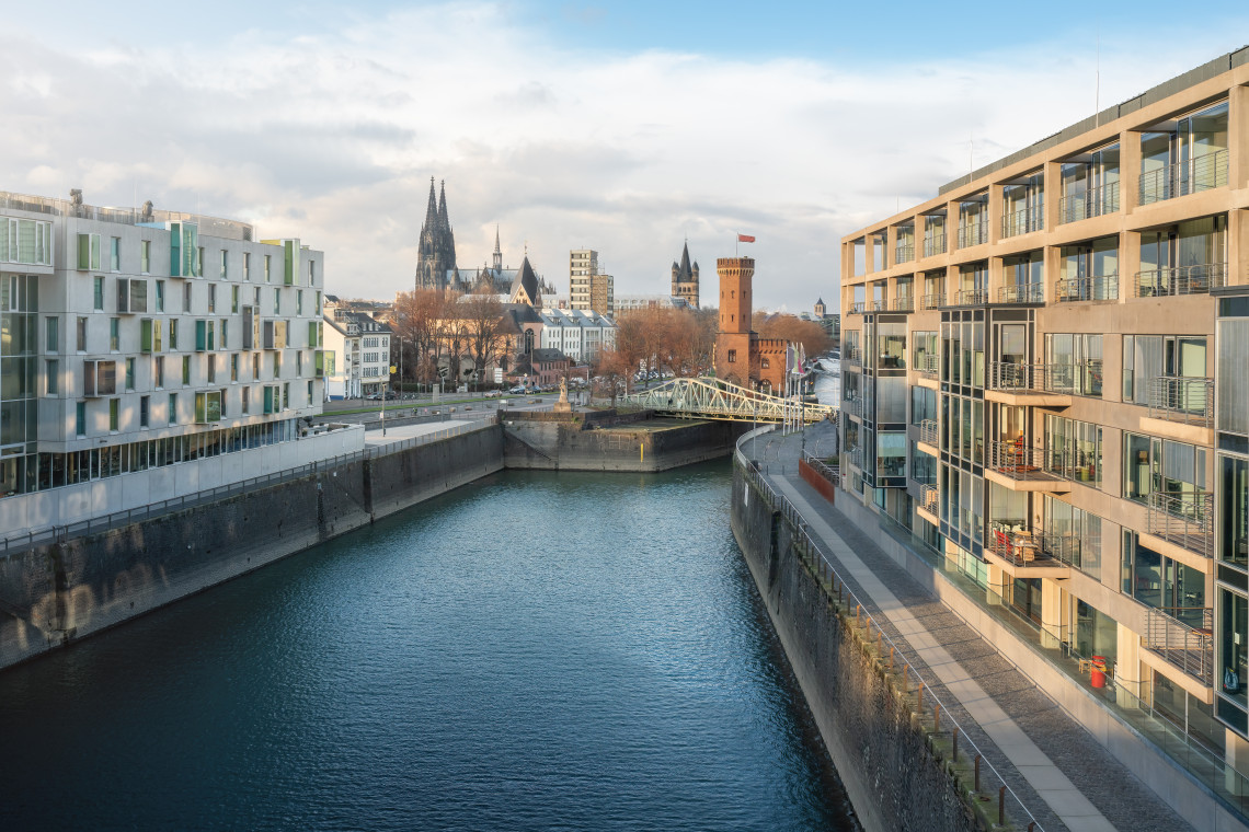 Modern waterfront buildings line the Rheinauhafen district in Cologne, with historic church towers and Cologne Cathedral visible in the distance.