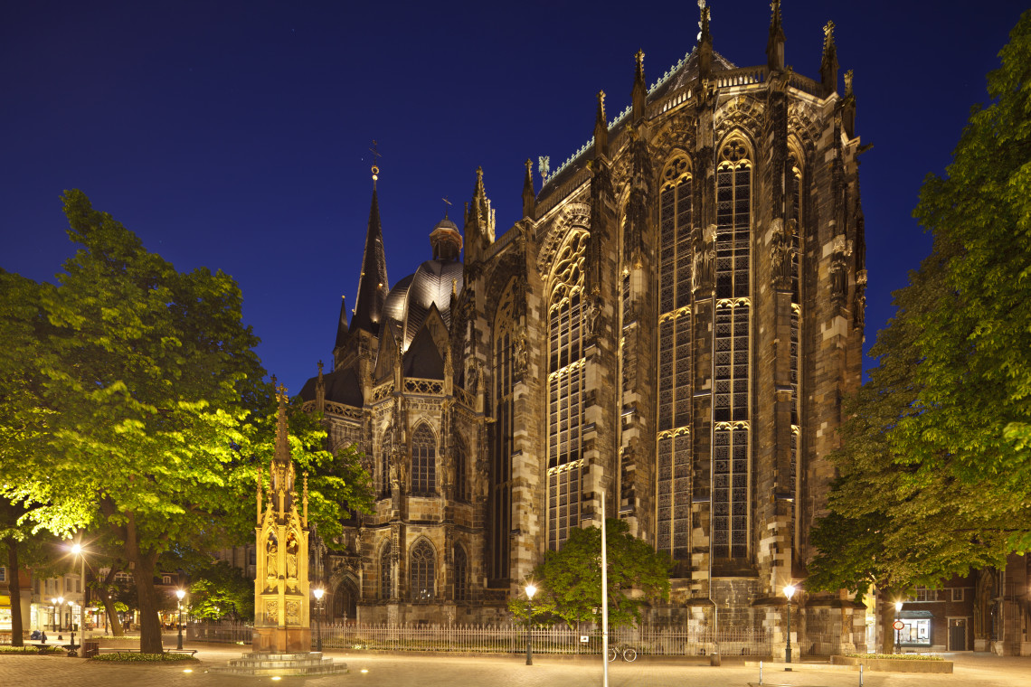 Illuminated Gothic architecture of Aachen Cathedral at night, framed by trees and historic monuments in central Aachen, Germany.