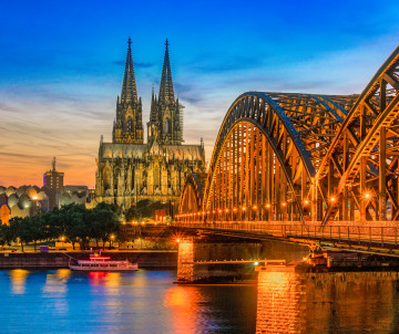 Cologne Cathedral rises beside the illuminated Hohenzollern Bridge over the Rhine River at dusk in Cologne, Germany.