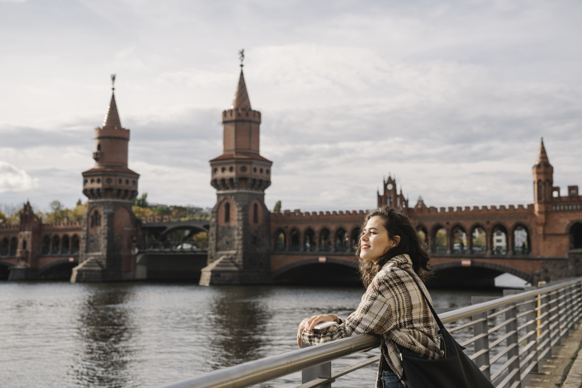 A woman rests against a riverside railing beside the Spree River in Berlin, Germany, with the red-brick towers and arches of Oberbaum Bridge behind her.