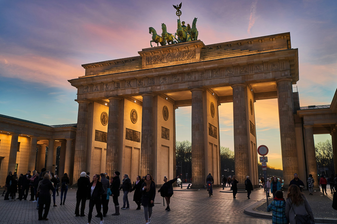 Visitors gather around the Brandenburg Gate in Berlin, Germany, as warm sunset light illuminates its neoclassical columns and the Quadriga sculpture above.