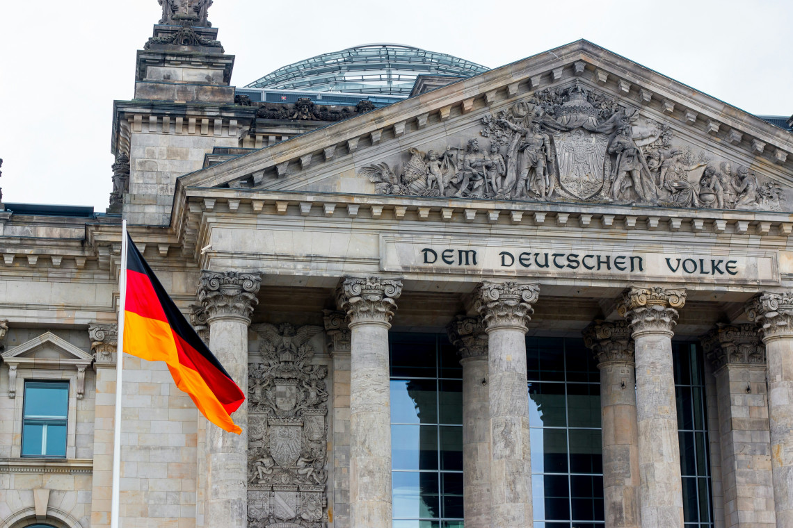 A German flag hangs in front of the Reichstag Building in Berlin, Germany. The detailed stone pediment, classical columns, &ldquo;Dem Deutschen Volke&rdquo; inscription and modern glass dome are visible.