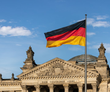 The German flag flies in front of the Reichstag Building in Berlin, Germany, with the historic stone façade and glass dome visible in the background.