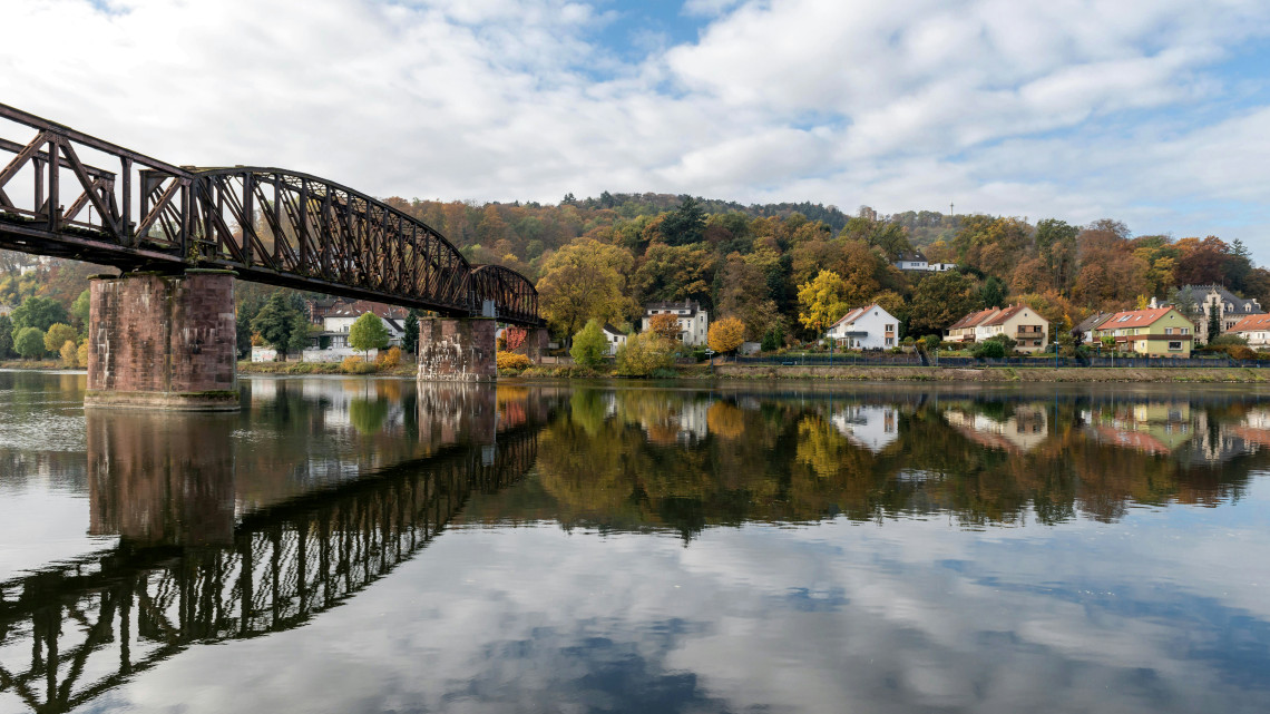 Dark steel truss bridge reflected in calm river water, with houses and wooded hills along the opposite bank in Hesse.