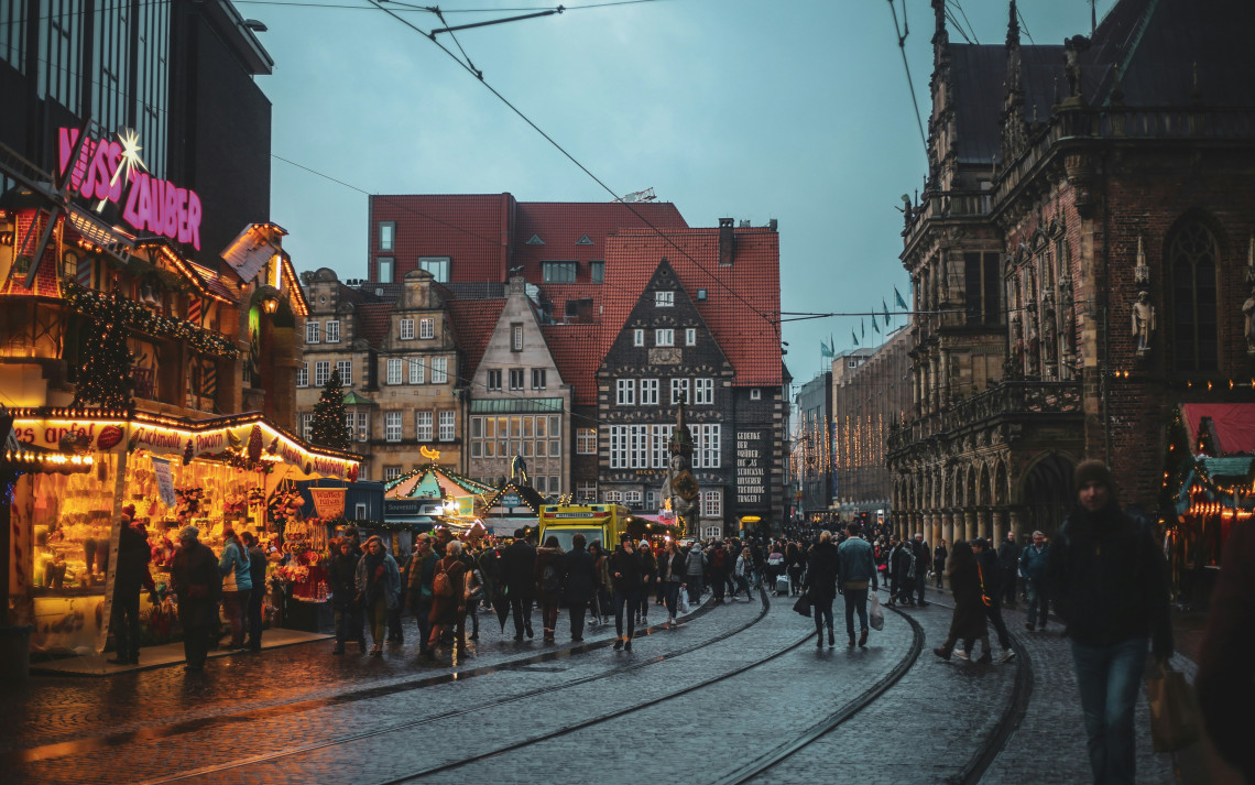 Crowded German Christmas market at dusk with illuminated wooden stalls, tram tracks and historic buildings lining the street.