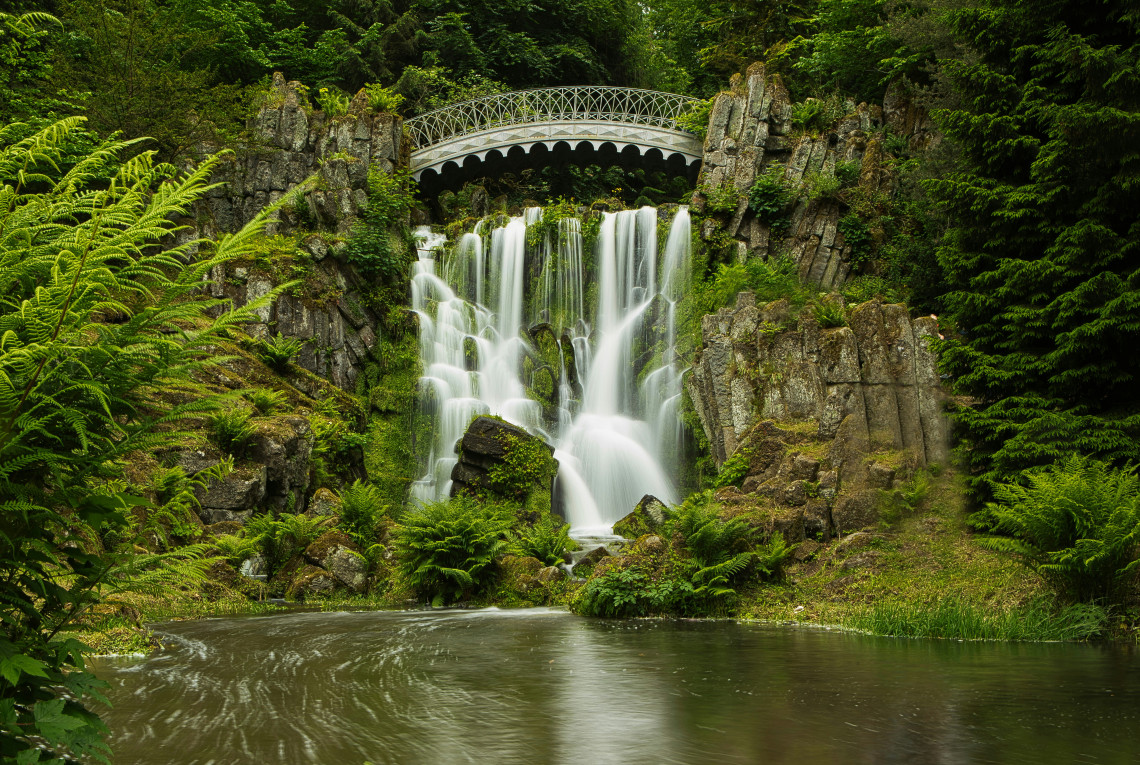 Water cascades beneath the decorative Teufelsbr&uuml;cke bridge into a forest pool surrounded by lush vegetation in Kassel.