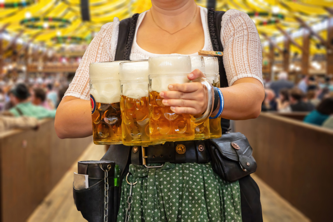 A server in traditional Bavarian dress carries several large beer steins through a busy Oktoberfest beer tent in Munich.