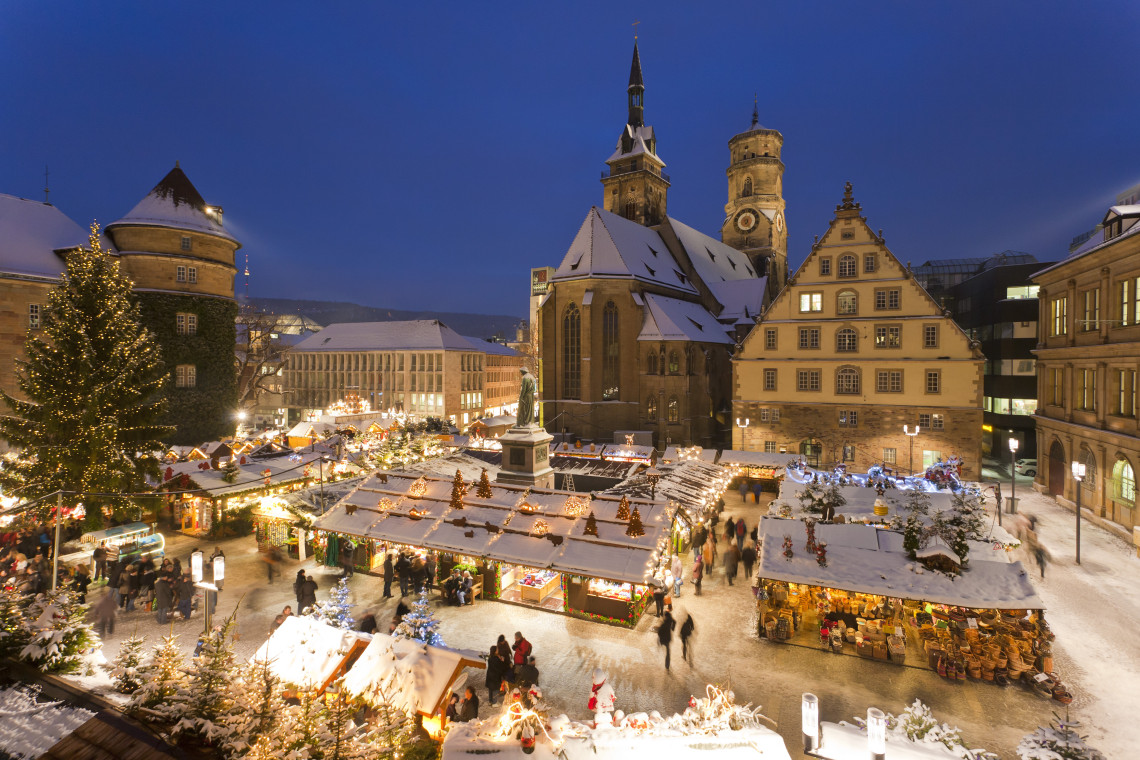 Snow-covered Christmas market stalls glow around Schillerplatz in Stuttgart, with historic buildings and the Stiftskirche rising behind them.