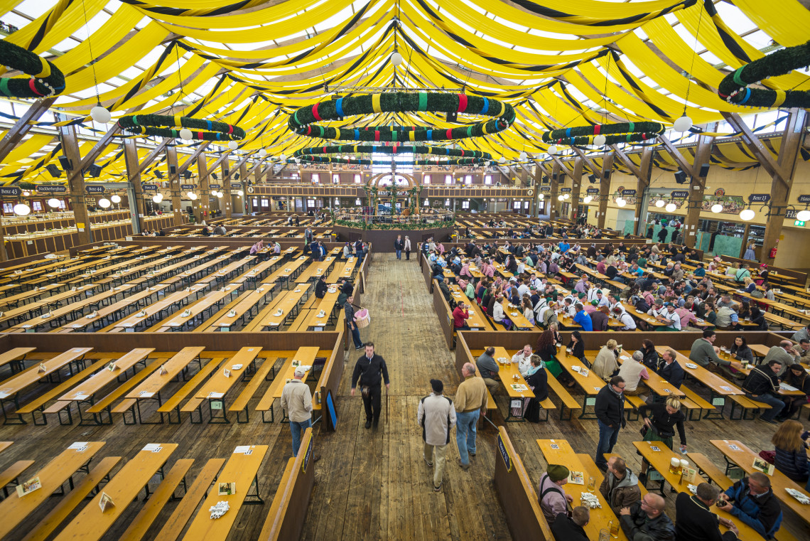 Long communal tables fill a traditional Oktoberfest beer hall in Munich beneath yellow decorations and large hanging wreaths.