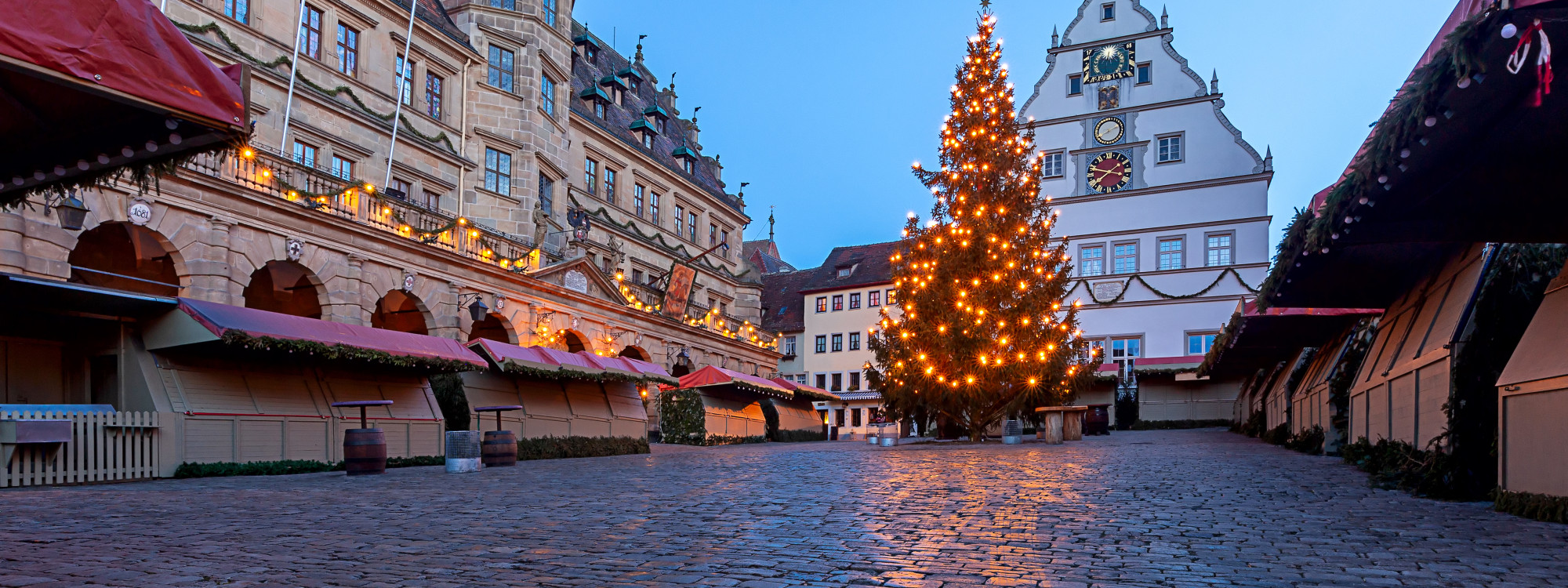 Early evening view of the Reiterlesmarkt Christmas market at Marktplatz in Rothenburg ob der Tauber, Germany. An illuminated Christmas tree stands between traditional market stalls, with the historic Ratstrinkstube and its clock overlooking the cobbleston